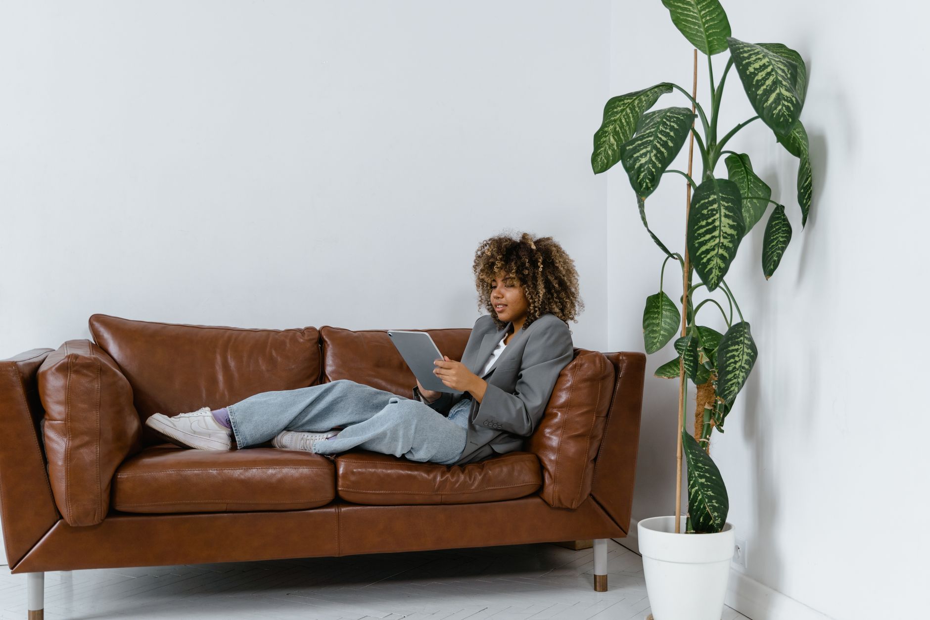 Woman lounging on a leather sofa with a tablet at home, next to a potted plant.